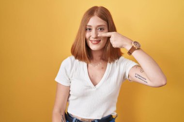 Sarı arka planda duran genç, kızıl saçlı bir kadın, el parmağı ve burunla işaret ediyor, neşeli bir şekilde gülümsüyor. güzellik konsepti 