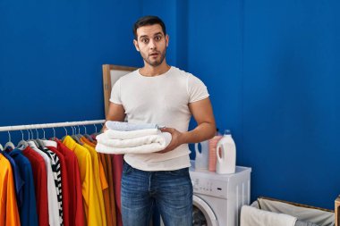 Young hispanic man with beard holding folded clean towels at laundry room afraid and shocked with surprise and amazed expression, fear and excited face. 