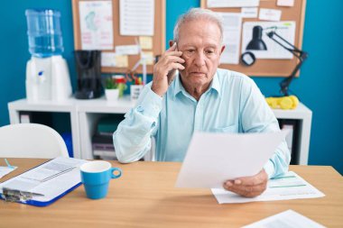 Middle age grey-haired man business worker talking on smartphone reading document at office