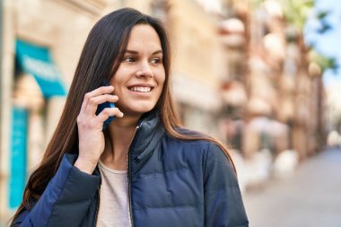 Genç, güzel, İspanyol bir kadın gülümsüyor ve caddede akıllı telefondan konuşuyor.