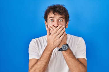 Mavi arka planda duran Latin kökenli genç adam yanlışlıkla elleri ile ağzını kapatırken şok oldu. gizli kavram. 