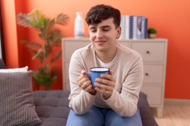 Non binary man drinking coffee sitting on sofa at home
