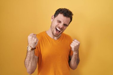 Young hispanic man standing over yellow background very happy and excited doing winner gesture with arms raised, smiling and screaming for success. celebration concept. 