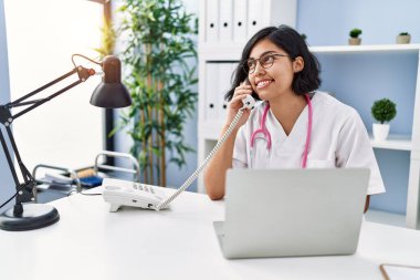 Doktor üniforması giyen genç Latin kadın klinikte telefonla konuşuyor.