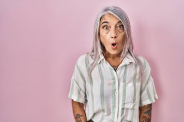 Pembe arka planda duran gri saçlı, ağzı açık, korkmuş ve şaşırmış bir orta yaşlı kadın. 