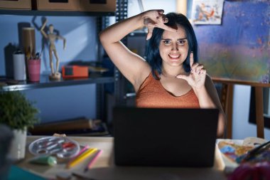 Young modern girl with blue hair sitting at art studio with laptop at night smiling making frame with hands and fingers with happy face. creativity and photography concept. 