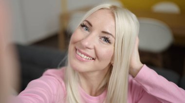 Genç sarışın kadın evdeki kanepede kamerayla selfie çekiyor.