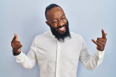 African american man standing over blue background shouting with crazy expression doing rock symbol with hands up. music star. heavy music concept. 