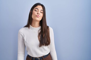 Young brunette woman standing over blue background looking at the camera blowing a kiss on air being lovely and sexy. love expression. 