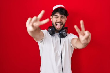 Hispanic man with beard wearing gamer hat and headphones smiling with tongue out showing fingers of both hands doing victory sign. number two. 