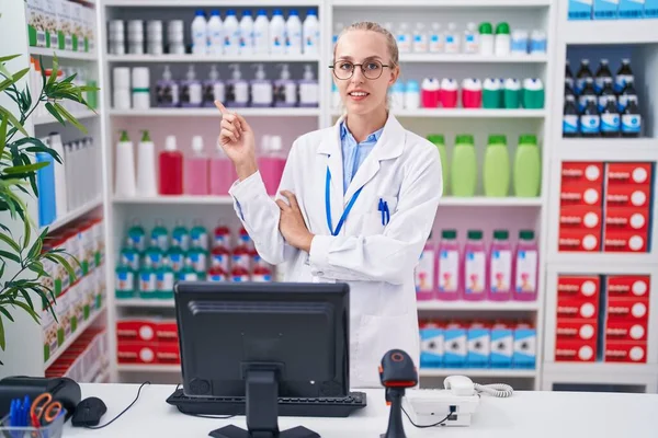 Young caucasian woman working at pharmacy drugstore with a big smile on face, pointing with hand and finger to the side looking at the camera.