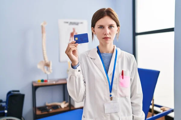 Young brunette woman working at pain recovery clinic holding credit card thinking attitude and sober expression looking self confident