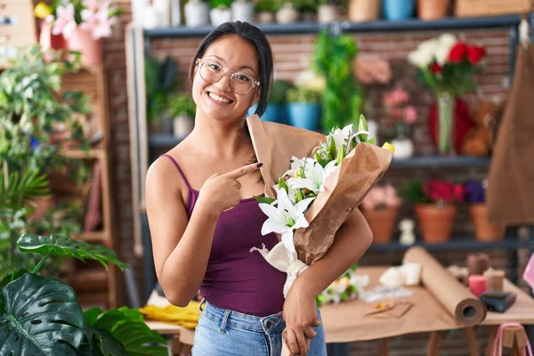 Asian young woman at florist shop holding bouquet of flowers smiling happy pointing with hand and finger