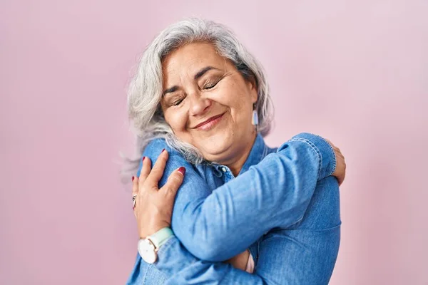 Middle age woman with grey hair standing over pink background hugging oneself happy and positive, smiling confident. self love and self care