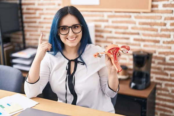 Young girl with blue hair holding model of female genital organ at the office complaining for menstruation pain surprised with an idea or question pointing finger with happy face, number one