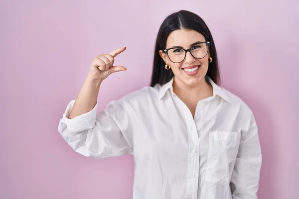 Young brunette woman standing over pink background smiling and confident gesturing with hand doing small size sign with fingers looking and the camera. measure concept.