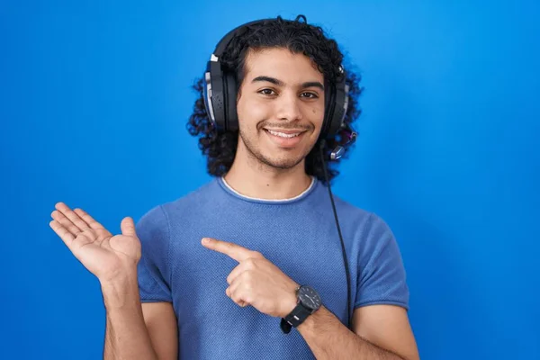 西班牙裔男人 一边用耳机听音乐 一边用手拿着相机 一边用手指指指 一边惊讶地微笑着 — 图库照片