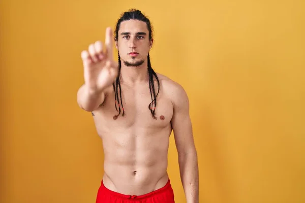 Hispanic man with long hair standing shirtless over yellow background pointing with finger up and angry expression, showing no gesture