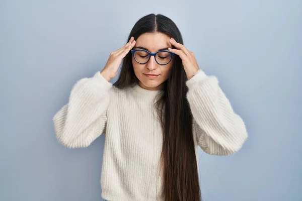 ストレスが原因で頭の痛みのために頭の上に手で青の背景にカジュアルなセーターを身に着けている若いヒスパニック系の女性 片頭痛 — ストック写真