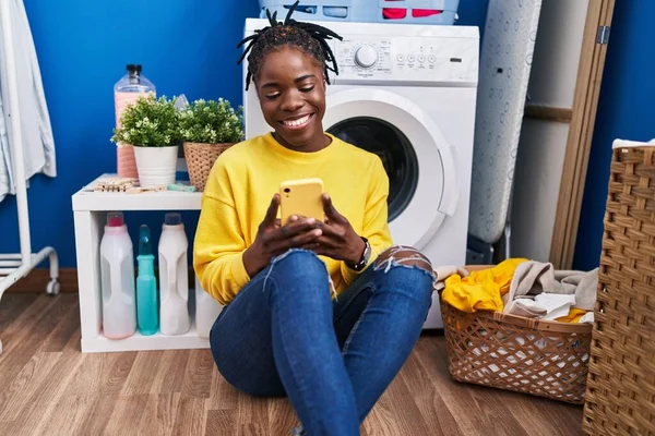 African american woman using smartphone waiting for washing machine at laundry room