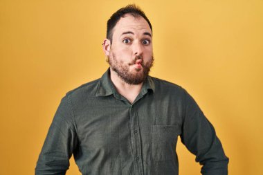 Plus size hispanic man with beard standing over yellow background making fish face with lips, crazy and comical gesture. funny expression. 