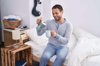 Young man drinking cup of coffee sitting on bed at bedroom