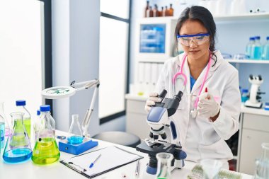 Young chinese woman wearing scientist uniform using microscope at laboratory