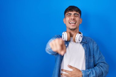 Young hispanic man standing over blue background laughing at you, pointing finger to the camera with hand over body, shame expression 