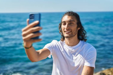 Genç İspanyol adam sahilde kayanın üzerinde otururken akıllı telefonuyla selfie çekiyor.