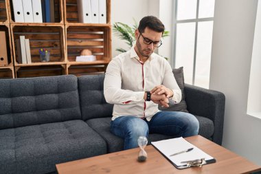 Young hispanic man with beard working at consultation office checking the time on wrist watch, relaxed and confident 