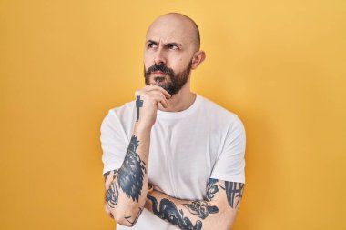 Sarı arka planda elleri çenesinde dikilen ve soru sormayı düşünen Latin kökenli genç bir adam, düşünceli bir ifade. Düşünceli bir yüzle gülümsüyor. şüphe kavramı. 