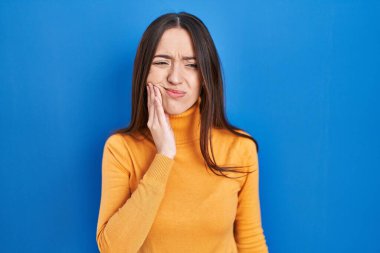 Mavi arka planda dikilen esmer kadın diş ağrısı ya da diş ağrısı yüzünden elleriyle dudaklarına dokunuyor. dişçi 