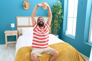 Young hispanic man waking up stretching arms at bedroom