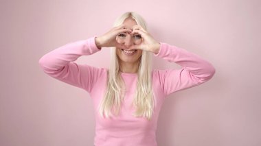 Young blonde woman smiling confident doing heart gesture with hands over isolated pink background