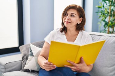 Orta yaşlı kadın kitap okuyor. Evdeki kanepede oturuyor.