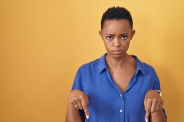 Sarı arka plan üzerinde duran Afrikalı Amerikalı kadın üzgün ve üzgün görünüyor, parmaklarıyla yön gösteriyor, mutsuz ve depresif.. 