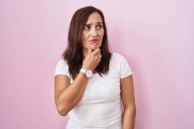 Middle age brunette woman standing over pink background thinking worried about a question, concerned and nervous with hand on chin 