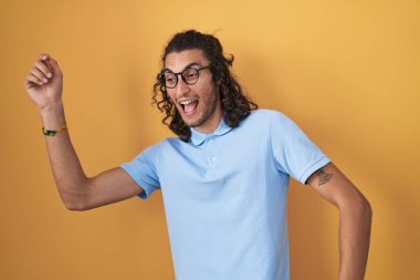 Young hispanic man standing over yellow background dancing happy and cheerful, smiling moving casual and confident listening to music 