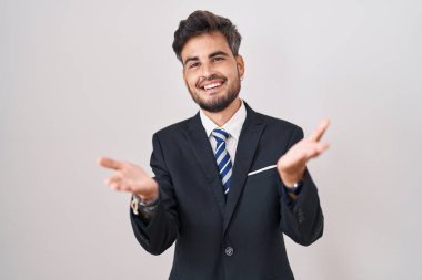 Young hispanic man with tattoos wearing business suit and tie smiling cheerful offering hands giving assistance and acceptance. 