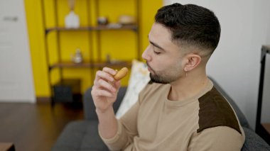 Genç Arap adam evdeki kanepede oturmuş donut yiyor.