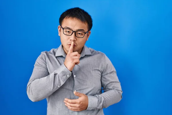 Young chinese man standing over blue background asking to be quiet with finger on lips. silence and secret concept.