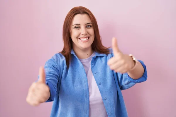 Young hispanic woman with red hair standing over pink background approving doing positive gesture with hand, thumbs up smiling and happy for success. winner gesture.