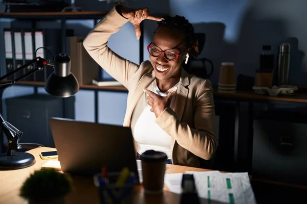 Beautiful black woman working at the office at night smiling making frame with hands and fingers with happy face. creativity and photography concept.