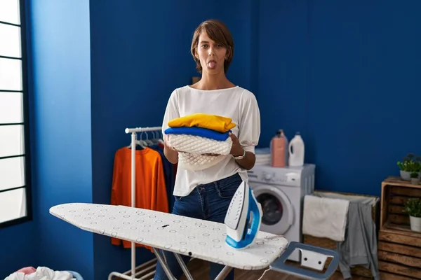 Brunette woman holding folded laundry after ironing sticking tongue out happy with funny expression.