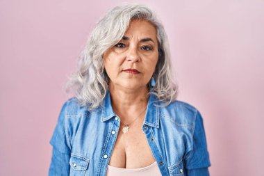 Middle age woman with grey hair standing over pink background relaxed with serious expression on face. simple and natural looking at the camera. 