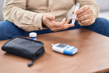 Orta yaşlı, kır saçlı, glukoz ölçen adam evdeki kanepede oturuyor.