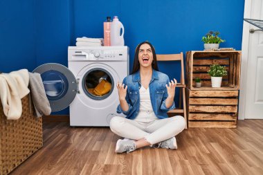 Hispanic woman doing laundry sitting on the floor crazy and mad shouting and yelling with aggressive expression and arms raised. frustration concept. 