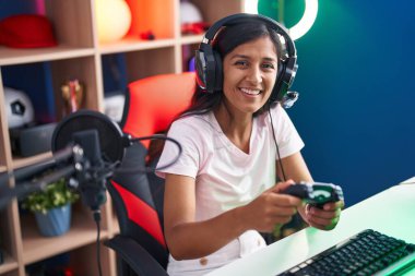 Young beautiful hispanic woman streamer playing video game using joystick at gaming room