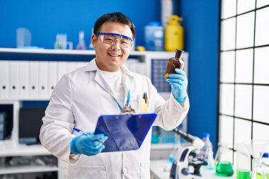 Young chinese man wearing scientist uniform reading clipboard at laboratory