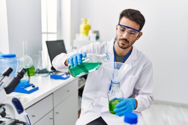 Young hispanic man wearing scientist uniform pouring liquid on test bottle at laboratory